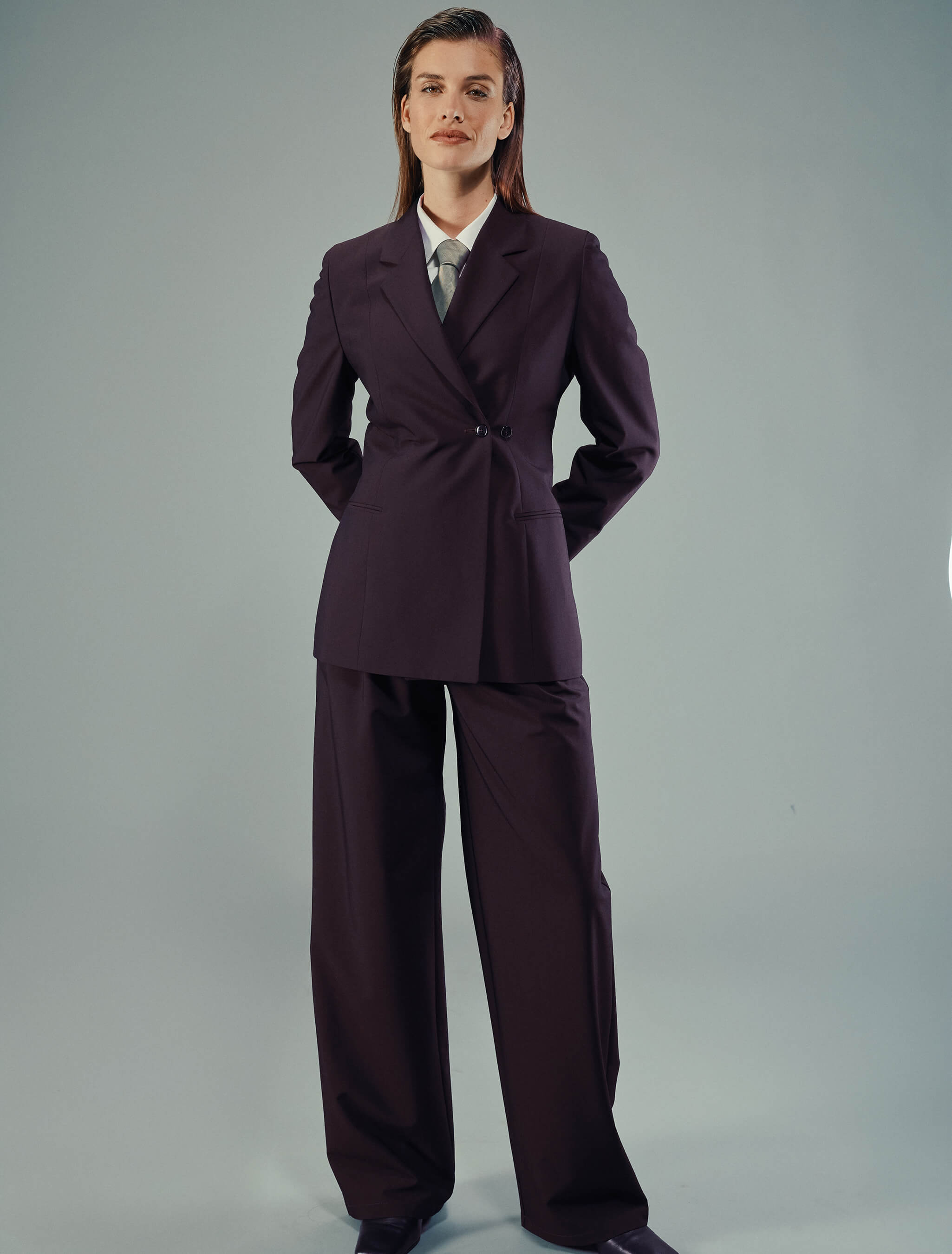 En kvinne har på seg en Women’s Avira Blazer i fargen Burgundy Berry, en hvit skjorte, et grått slips og matchende bukser med bredt ben og dobbeltspent.